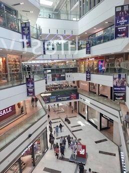 A multi-level shopping mall with large glass railings and visible store fronts including fashion retailers. People are walking throughout the space, with some carrying shopping bags. Sales banners are prominently displayed.