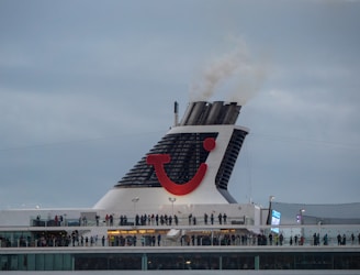 A large cruise ship chimney with a distinctive red and blue logo emits smoke into a cloudy sky. Below, many people stand on the ship's deck, suggesting observation or social activity.