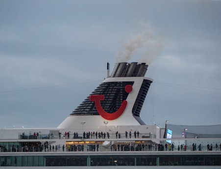 A large cruise ship chimney with a distinctive red and blue logo emits smoke into a cloudy sky. Below, many people stand on the ship's deck, suggesting observation or social activity.