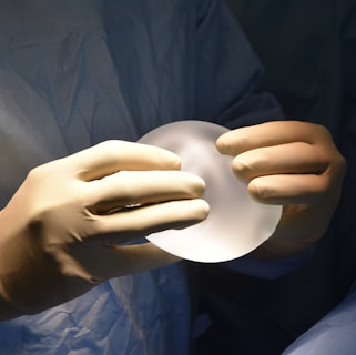 A pair of hands wearing surgical gloves holds a translucent, round, gel-like object, likely a medical implant. The background features a fabric that resembles surgical scrubs.
