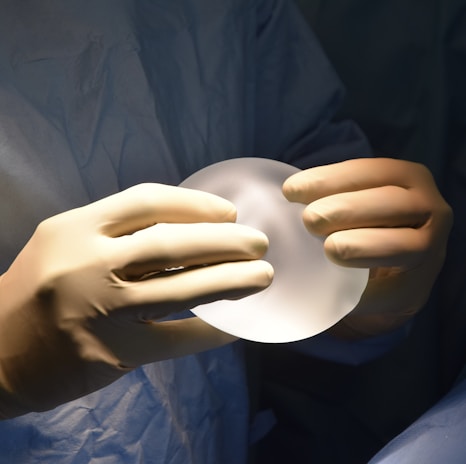 A pair of hands wearing surgical gloves holds a translucent, round, gel-like object, likely a medical implant. The background features a fabric that resembles surgical scrubs.
