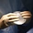 A pair of hands wearing surgical gloves holds a translucent, round, gel-like object, likely a medical implant. The background features a fabric that resembles surgical scrubs.