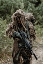 A person dressed in camouflage gear, including a ghillie suit, is holding a rifle and moving through a dense forest. The surroundings are lush with greenery and natural foliage.