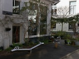 An outdoor urban space with playground equipment, surrounded by buildings with white walls and large windows. Plants and bushes are placed in pots around the area, creating a balance between nature and architecture. The surface is paved, and a street sign is visible on one of the building walls.