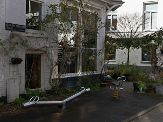 An outdoor urban space with playground equipment, surrounded by buildings with white walls and large windows. Plants and bushes are placed in pots around the area, creating a balance between nature and architecture. The surface is paved, and a street sign is visible on one of the building walls.