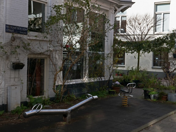 An outdoor urban space with playground equipment, surrounded by buildings with white walls and large windows. Plants and bushes are placed in pots around the area, creating a balance between nature and architecture. The surface is paved, and a street sign is visible on one of the building walls.