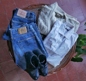A neatly arranged collection of clothing items includes a pair of blue denim jeans with leather patches, a cream-colored knit sweater, a crisp white dress shirt, and a pair of black leather shoes. These items are laid out on a wooden surface with a red tiled floor and green plant leaves visible in the background.