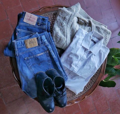 A neatly arranged collection of clothing items includes a pair of blue denim jeans with leather patches, a cream-colored knit sweater, a crisp white dress shirt, and a pair of black leather shoes. These items are laid out on a wooden surface with a red tiled floor and green plant leaves visible in the background.
