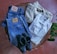 A neatly arranged collection of clothing items includes a pair of blue denim jeans with leather patches, a cream-colored knit sweater, a crisp white dress shirt, and a pair of black leather shoes. These items are laid out on a wooden surface with a red tiled floor and green plant leaves visible in the background.