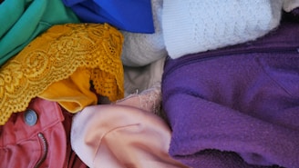 Various fabrics of different colors and textures are piled together. The materials include green, blue, white, purple, yellow, pink, and red clothing items, some of which feature buttons, zippers, and lace details.