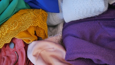 Various fabrics of different colors and textures are piled together. The materials include green, blue, white, purple, yellow, pink, and red clothing items, some of which feature buttons, zippers, and lace details.
