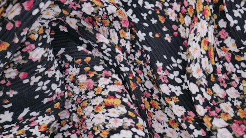 A close-up view of a patterned fabric featuring small floral designs in pink, white, and yellow against a black background. The fabric appears to have a textured and slightly wrinkled surface.