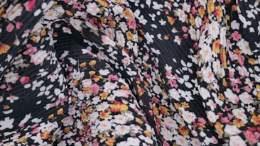 A close-up view of a patterned fabric featuring small floral designs in pink, white, and yellow against a black background. The fabric appears to have a textured and slightly wrinkled surface.