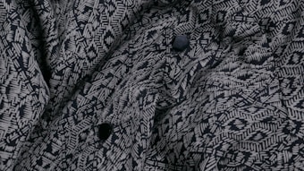 A close-up view of a fabric with a complex geometric pattern consisting of white lines and shapes on a dark background. The fabric appears to have a textured surface with multiple folds and several black buttons are visible.