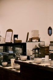A cozy café interior with a wooden counter displaying a variety of cups and glassware. Behind the counter, there are items like a display case, menu board, and stacked paper cups. The background features soft lighting and a simple, minimalistic decor, contributing to a relaxed atmosphere.