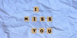 A message spelled out with wooden letter tiles on a crumpled, light blue paper background reads 'I MISS YOU'. The tiles form a neat pyramid-like arrangement with 'I' at the top, 'MISS' in the middle, and 'YOU' at the bottom.