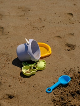 Various colorful beach toys, including a white bucket, yellow sieve, green molds, and a blue scoop, are scattered on sandy beach terrain. The sand displays footprints and small pebbles.
