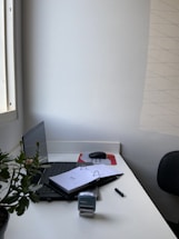 A tidy office desk with a laptop partially closed, a potted plant on the left, and an open binder with papers. A stamp, pen, mouse, and mousepad are also visible on the white desk. The desk is placed against a white wall, with a window to the side.