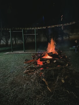 A campfire is burning brightly in a dark outdoor setting, surrounded by grass and a horizontal string of colorful flags hanging nearby. A playground structure is visible in the background, and a person sits on a bench close by.