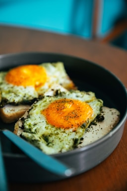 Two slices of bread topped with fried eggs, seasoned with herbs, and served in a round pan. The yolks are bright yellow and slightly runny, complemented by a sprinkle of black pepper and herbs. A teal handle of a utensil is visible, indicating a kitchen or dining setting.