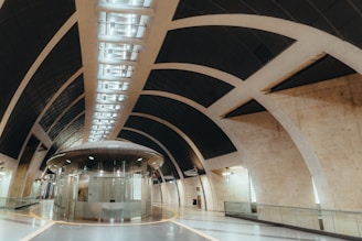A modern architectural space featuring large arched ceilings with a grid pattern and a central cylindrical structure made of glass and metallic materials. The space appears open and is well-lit with overhead lights. The flooring is smooth and has a circular design that complements the architecture.