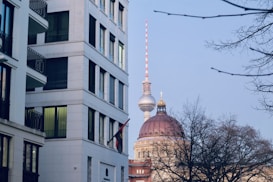 A modern building with white walls and balconies is on the left, while in the background, the Berlin TV Tower and a domed building with a golden cross on top are visible. The scene is framed by bare trees and the sky is clear.