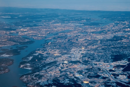 An aerial view of a large urban area with a network of roads and buildings. A river or a body of water winds through the city, and there are patches of greenery and land surrounding the water. The scene is expansive, showing a significant amount of development.