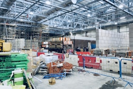 An indoor construction site area filled with materials such as stacks of pallets, various tools, protective barriers, and construction equipment. Scaffolding is visible in the background, and the space has a steel framework ceiling with large ducts. There are several stacks of pipes and cables, as well as safety signs throughout the area.