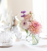 A small glass vase holds a bouquet of pink and purple flowers, including dahlias and roses. The vase is set on a white tablecloth, accompanied by elegant glassware. Soft lighting enhances the gentle and serene atmosphere.