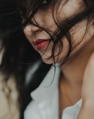 The close-up photograph captures a woman with long, dark hair partially covering her face. Her red lips and a hint of her eye are visible through the strands of hair. She wears a white garment, which adds a touch of contrast. The image focuses on the intricacy of hair and the softness of her skin.