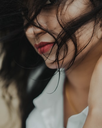 The close-up photograph captures a woman with long, dark hair partially covering her face. Her red lips and a hint of her eye are visible through the strands of hair. She wears a white garment, which adds a touch of contrast. The image focuses on the intricacy of hair and the softness of her skin.