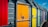 Brightly colored doors of beach huts in a row, featuring a variety of vibrant colors including red, yellow, orange, green, blue, and pink. Each door has a padlock and metal brace, and they are attached to grey slatted wooden walls under a blue sky with a few small clouds.