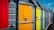 Brightly colored doors of beach huts in a row, featuring a variety of vibrant colors including red, yellow, orange, green, blue, and pink. Each door has a padlock and metal brace, and they are attached to grey slatted wooden walls under a blue sky with a few small clouds.