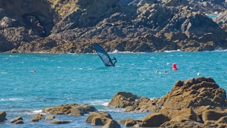 A rocky shoreline with turquoise blue water in the foreground. A windsurfer is navigating the waters, surrounded by a few swimmers near a red buoy. In the background, rugged cliffs rise from the sea.