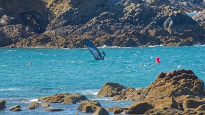 A rocky shoreline with turquoise blue water in the foreground. A windsurfer is navigating the waters, surrounded by a few swimmers near a red buoy. In the background, rugged cliffs rise from the sea.