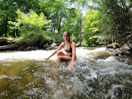 A serene woman sitting peacefully by a flowing stream surrounded by lush greenery, radiating calm vitality.