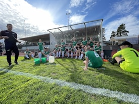 A group of athletes in green uniforms are taking a break near a sports field. They are seated and standing around a covered bench area. The sky is bright and partly cloudy, casting sunlight over the field.