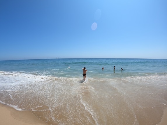 A sandy beach with gentle waves lapping at the shore. Several people are enjoying the water under a clear blue sky. The sun is bright, creating a warm and inviting atmosphere.