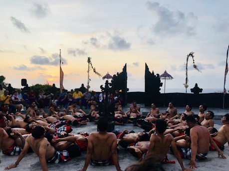 A group of shirtless men are sitting in a circle on an outdoor platform, surrounding a central point. They are engaged in a traditional Balinese dance performance at sunset. The background features an ornate stone structure and ceremonial decorations, with a few people watching from behind.
