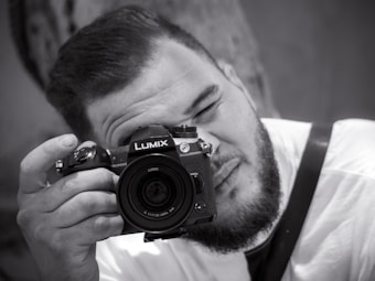A black and white image featuring a person with a beard holding a Lumix camera up to their face, seemingly taking a photo. The person is wearing a light-colored shirt and a strap is visible over their shoulder. The background is blurred, focusing attention on the individual and the camera.