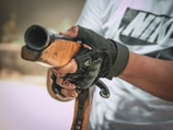 A person wearing a white shirt with black lettering is holding a wooden gun stock, with gloved hands gripping it firmly. The focus is on the hands and the gun, capturing the texture of the wood and the material of the gloves.