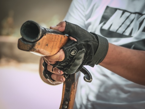 A person wearing a white shirt with black lettering is holding a wooden gun stock, with gloved hands gripping it firmly. The focus is on the hands and the gun, capturing the texture of the wood and the material of the gloves.