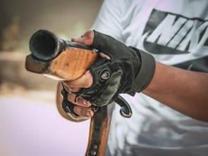 A person wearing a white shirt with black lettering is holding a wooden gun stock, with gloved hands gripping it firmly. The focus is on the hands and the gun, capturing the texture of the wood and the material of the gloves.