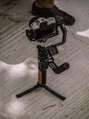 A camera mounted on a stabilizing gimbal is positioned on a concrete surface. A person wearing dark shoes stands nearby, partially visible in the background.