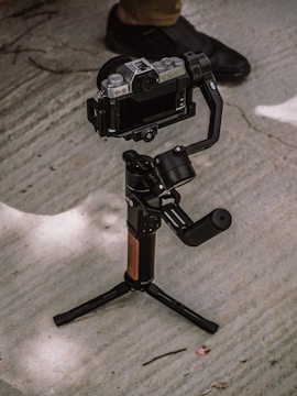A camera mounted on a stabilizing gimbal is positioned on a concrete surface. A person wearing dark shoes stands nearby, partially visible in the background.
