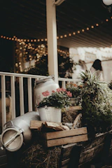 A rustic arrangement featuring metal milk churns, a crate with wooden logs, and vibrant flowers including red blossoms. The scene is set on a wooden cart with hay, and the background is softly lit with string lights that create a warm, bokeh effect. There is lush greenery surrounding the setup.