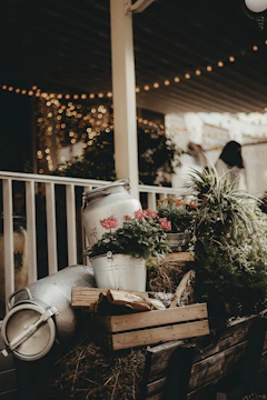 A rustic arrangement featuring metal milk churns, a crate with wooden logs, and vibrant flowers including red blossoms. The scene is set on a wooden cart with hay, and the background is softly lit with string lights that create a warm, bokeh effect. There is lush greenery surrounding the setup.