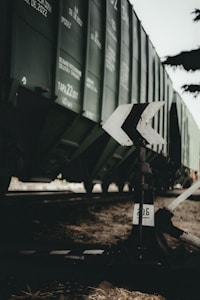 A close-up of a railway switch mechanism in front of a freight train. The train is green with visible text and numbers on its side. The setting appears to be an outdoor train yard, with earthy tones and scattered dry leaves on the ground.