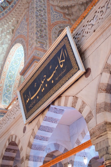 An intricately decorated interior with ornate arches and a prominently displayed rectangular sign featuring Arabic script. The architecture features detailed geometric and floral designs in muted colors, showcasing Islamic art and craftsmanship.