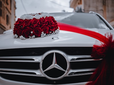 A white car is adorned with a decorative arrangement of vibrant red roses and white flowers on the hood. A red ribbon is elegantly draped across, enhancing the festive appearance. The emblem of the car manufacturer is prominently displayed on the front grille.